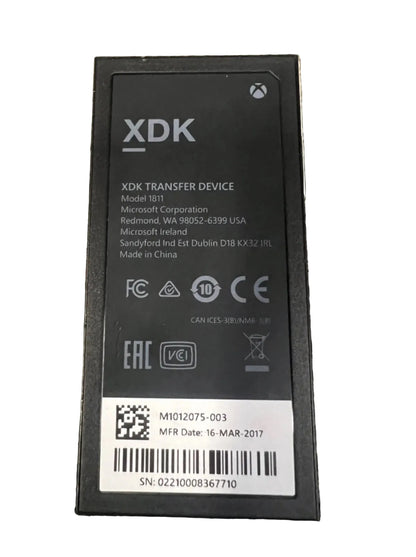 A black XDK transfer device battery or replacement cell with printed specifications, barcodes, and a white label, photographed on a textured beige carpet.